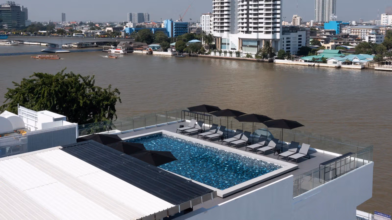 View down to a riverside rooftop pool, dark umbrellas and loungers arranged along the river