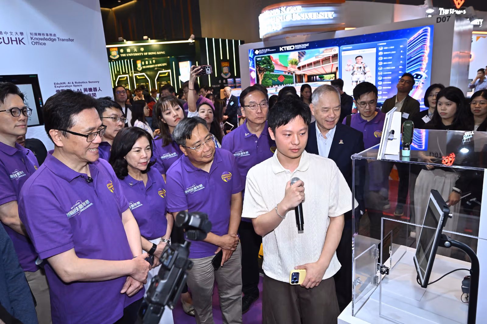 A presenter holding a microphone explains a display beside a transparent case and screens