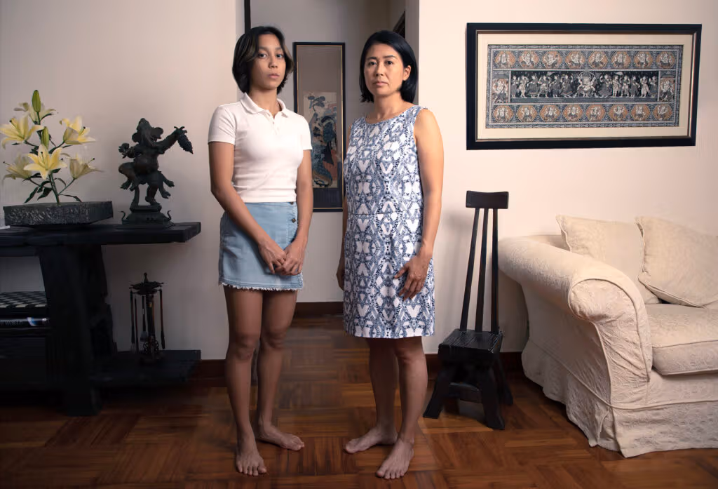 Two people stand barefoot at home beside a vase, sculpture, wall pictures, wooden chair and sofa