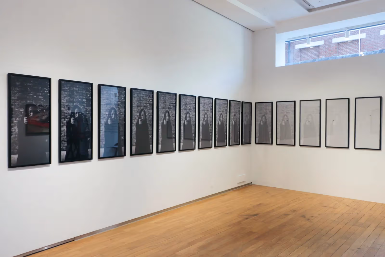 Vertically framed photographs form a sequence on white walls, with a figure and brick background shifting from dark to pale