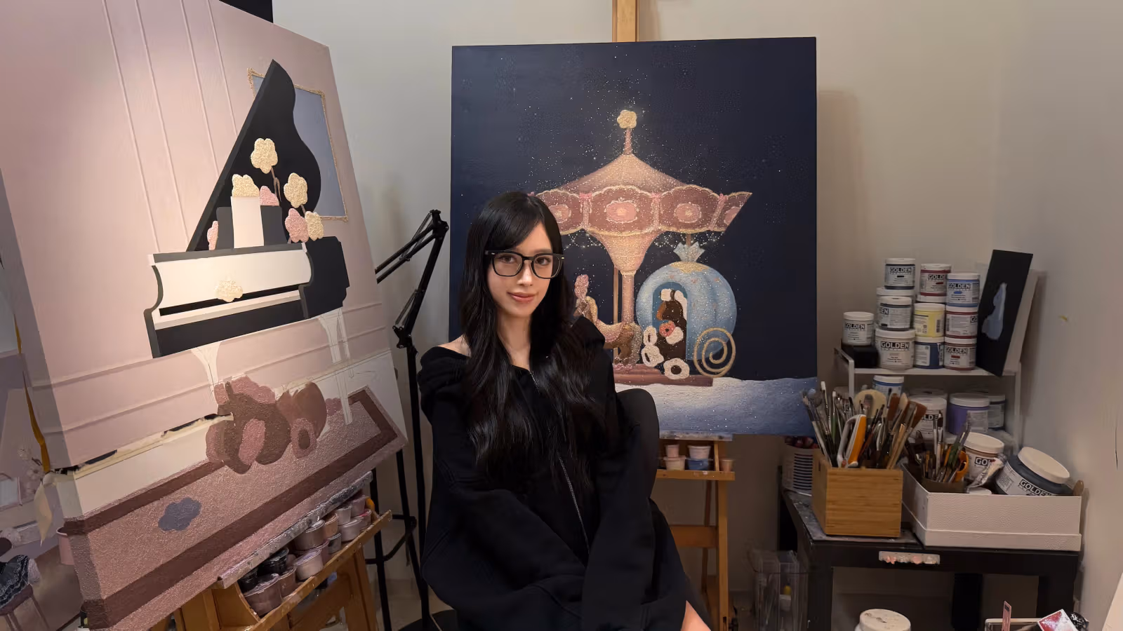 Esther Luo นั่งในสตูดิโอ มีผ้าใบทางซ้ายและด้านหลัง ทางขวาเรียงกระปุกสีกับพู่กัน