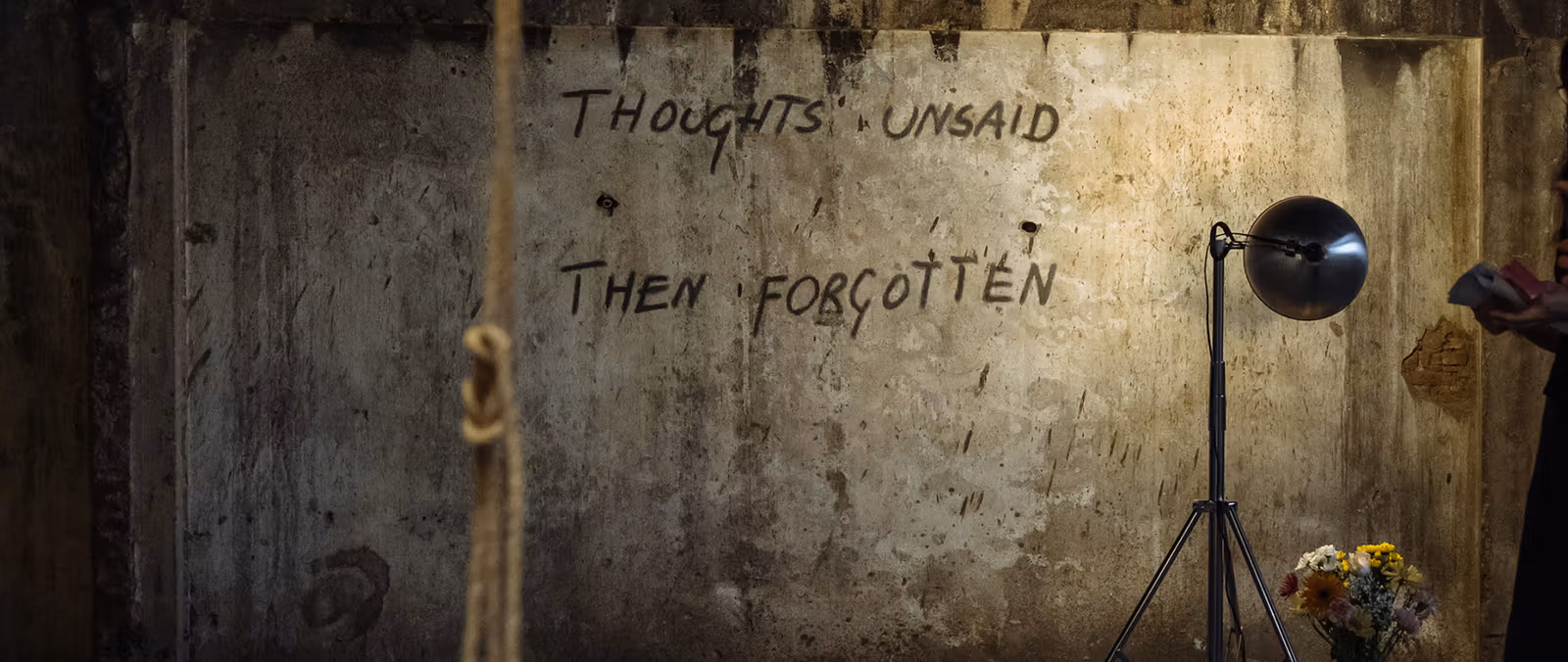 ผนังหยาบมีข้อความ Thoughts unsaid then forgotten โคมไฟขาตั้งสามขาส่องไปที่ข้อความ และเห็นช่อดอกไม้ด้านล่าง