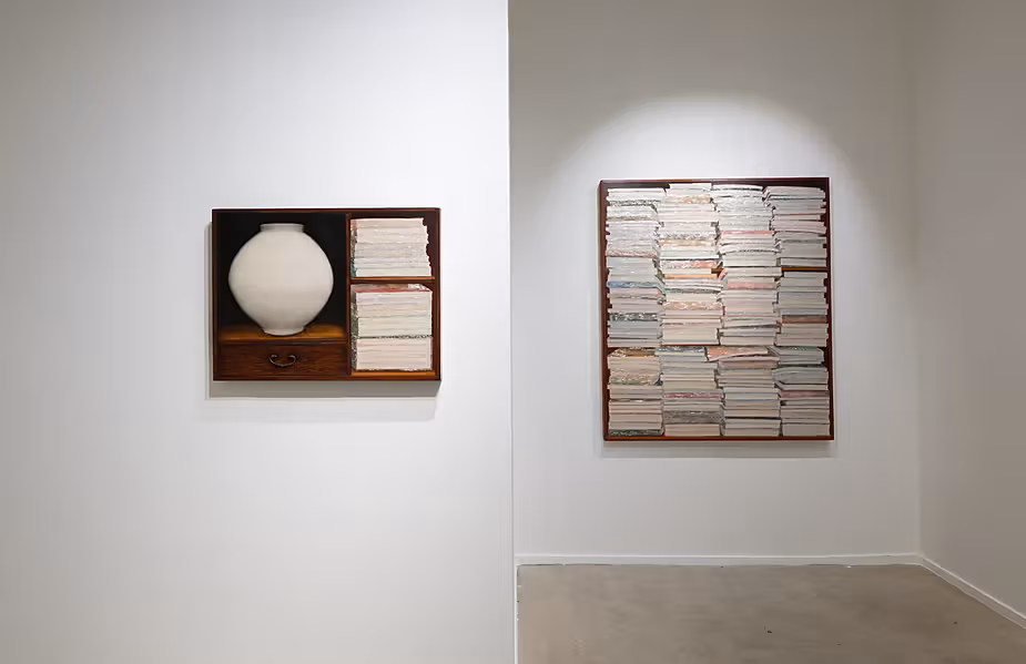 A white vessel and books in a wooden compartment at left; rows of white book spines fill the work at right.