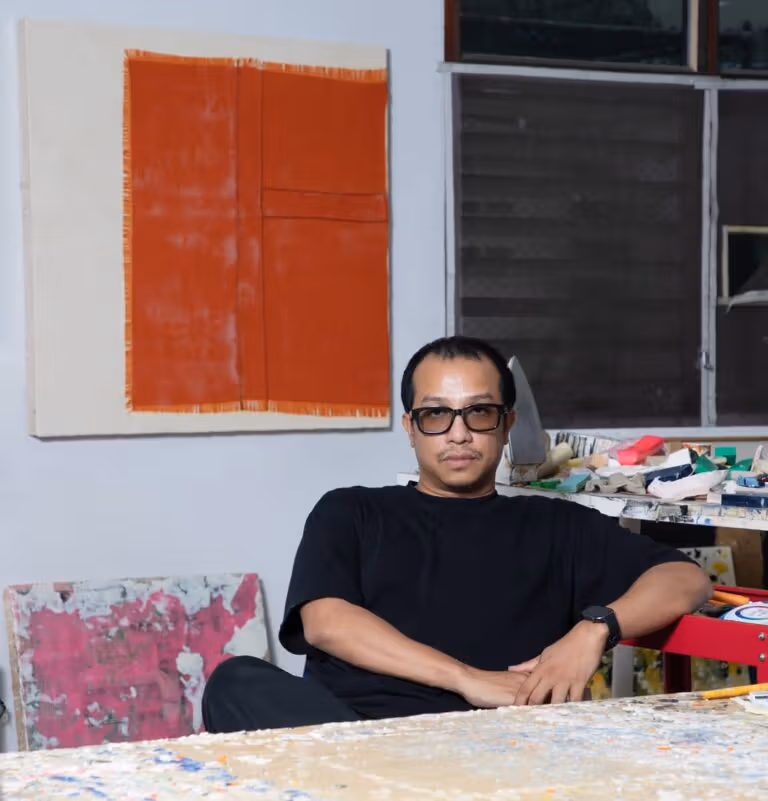 Artist Tada Hengsapkul, wearing glasses and a black shirt, seated in a working environment with artworks, tools and a paint-marked table