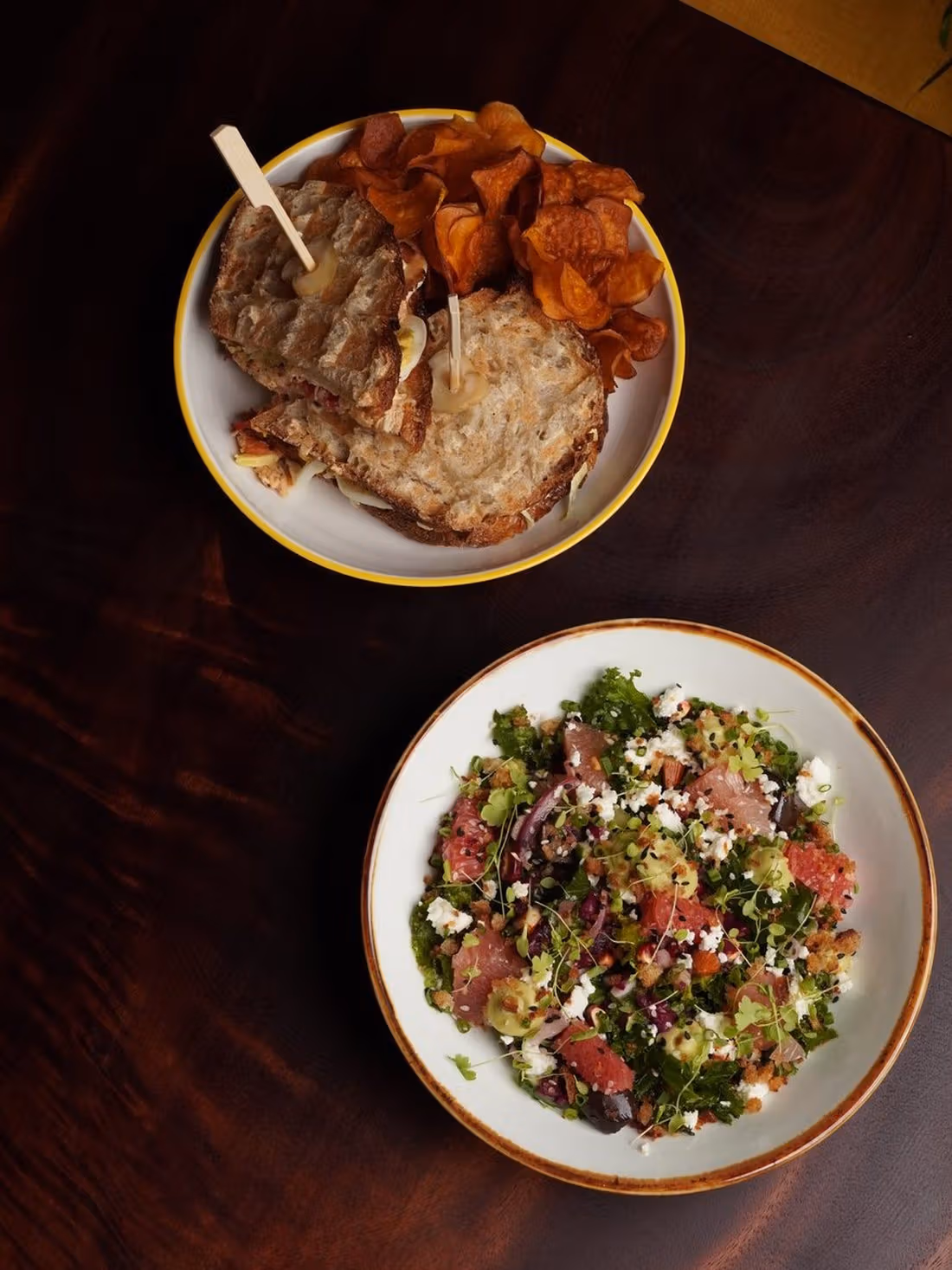 나무 테이블 위쪽 접시에 The Chicken Club 샌드위치와 고구마칩, 아래쪽 접시에 Kale Salad
