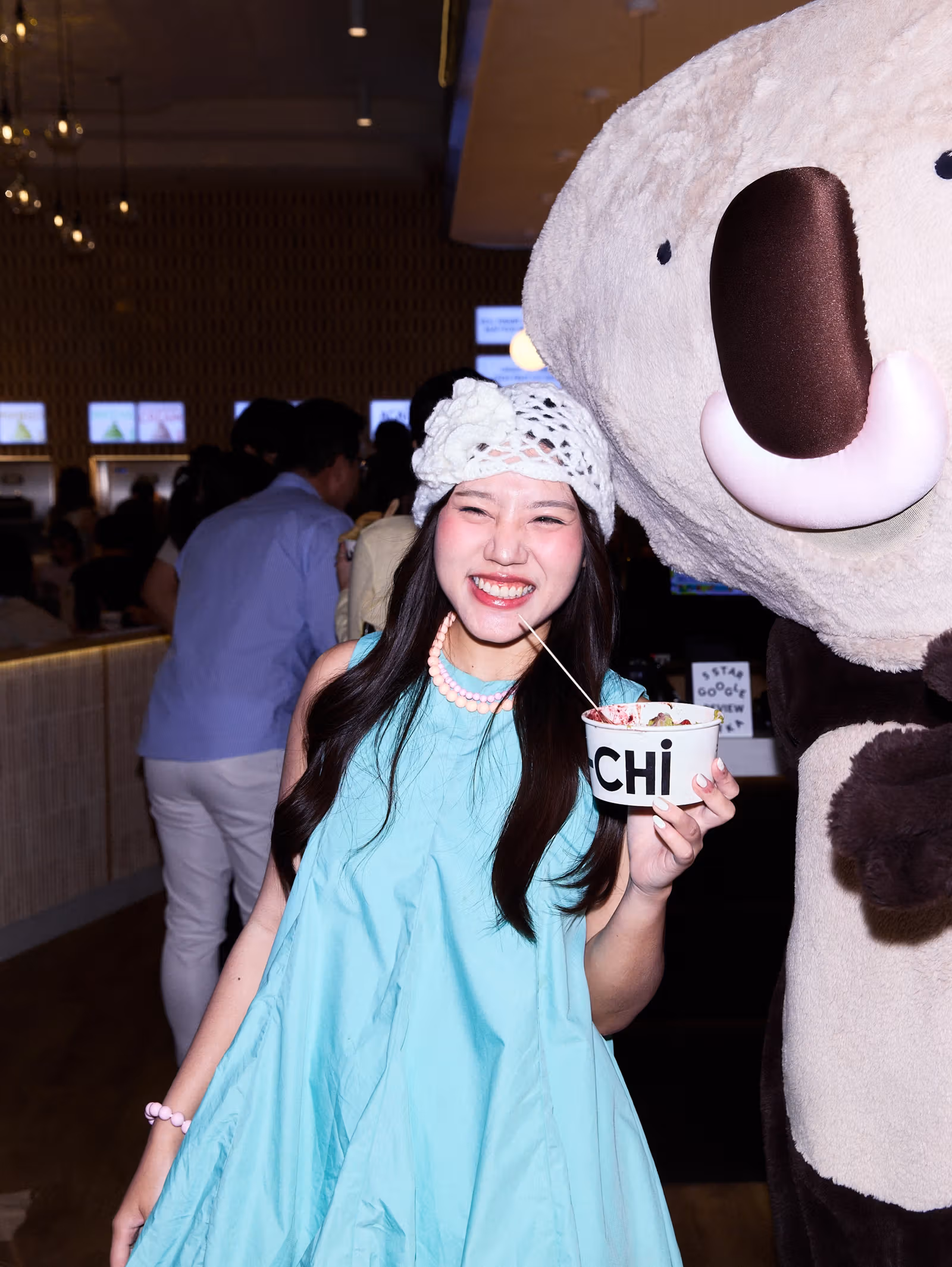 Yo-Chiカップを持つ人物と大きなコアラのキャラクター。背景に客とセルフサービス機が見える
