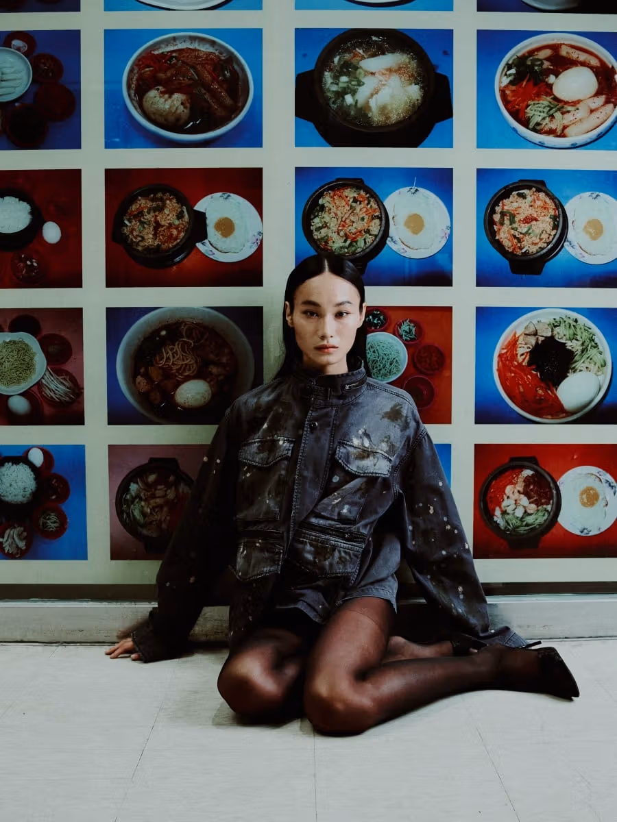 Model in a dark military jacket seated before a wall of food photographs