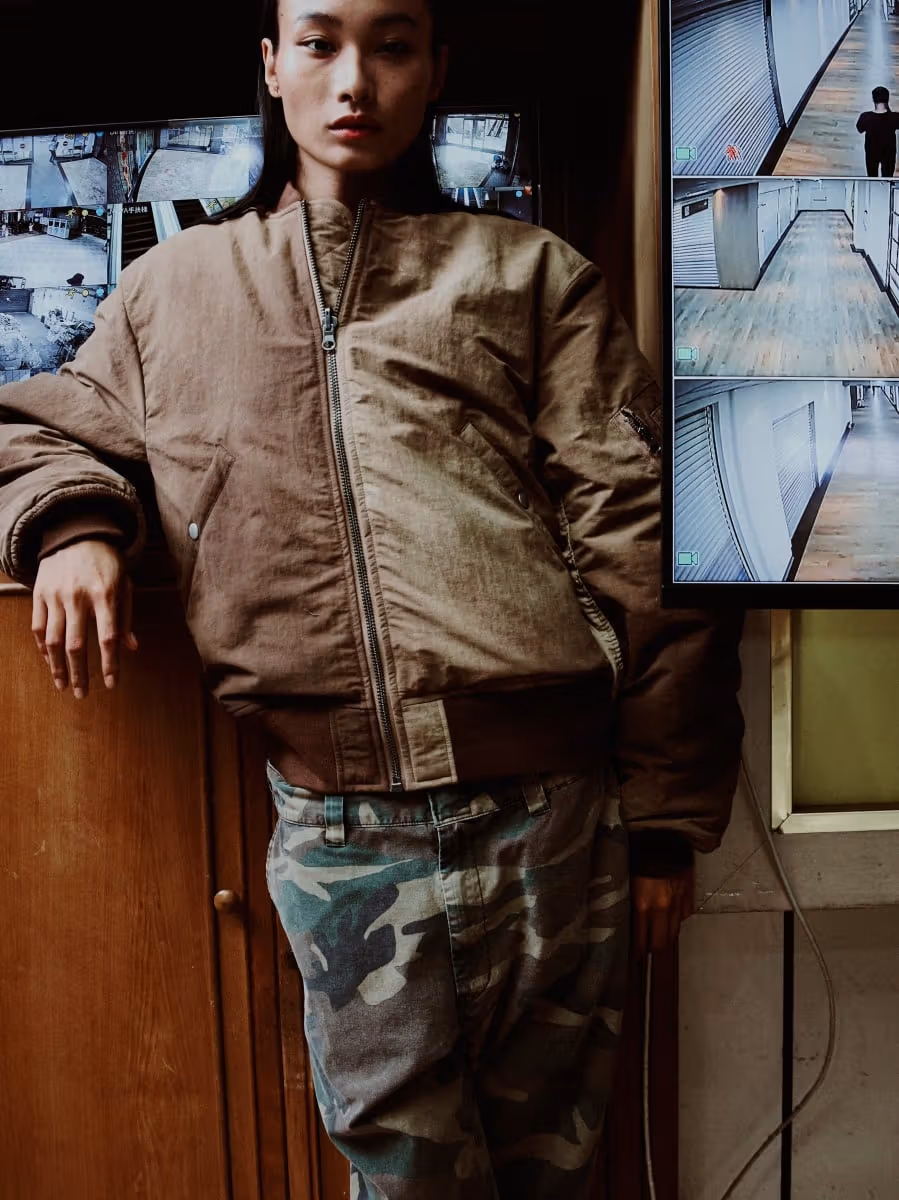 Model wearing a brown jacket and camouflage trousers beside surveillance screens