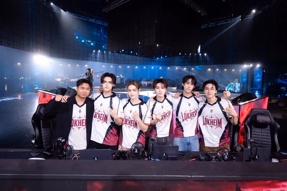 คนหกคนในเสื้อสีขาวแดงไวน์ยืนเรียงหลังโต๊ะแข่งขันสีแดง ด้านหน้ามีหูฟังและพัดลมเล็ก
