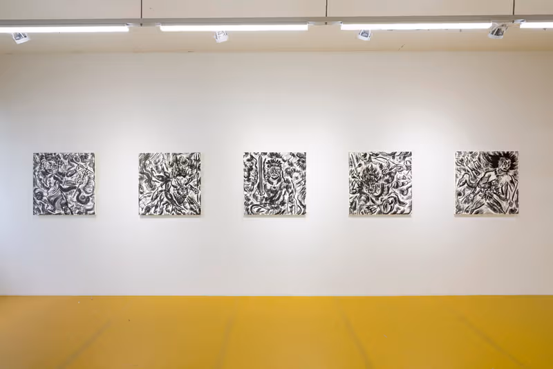 Five square black-and-white works hang along a white wall above a yellow gallery floor, with strip lighting overhead.