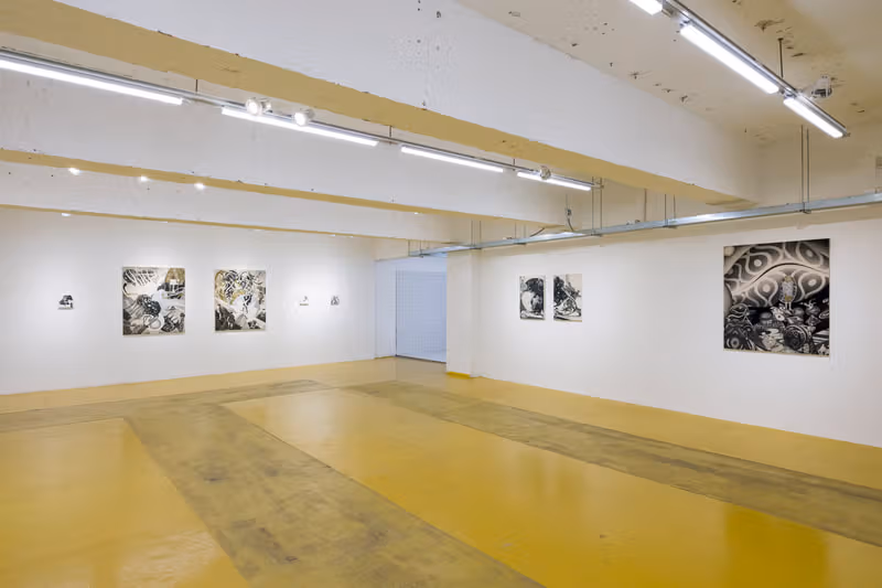 Black, grey and gold works of different sizes hang in a white-walled gallery with yellow floors, exposed beams and strip lights.