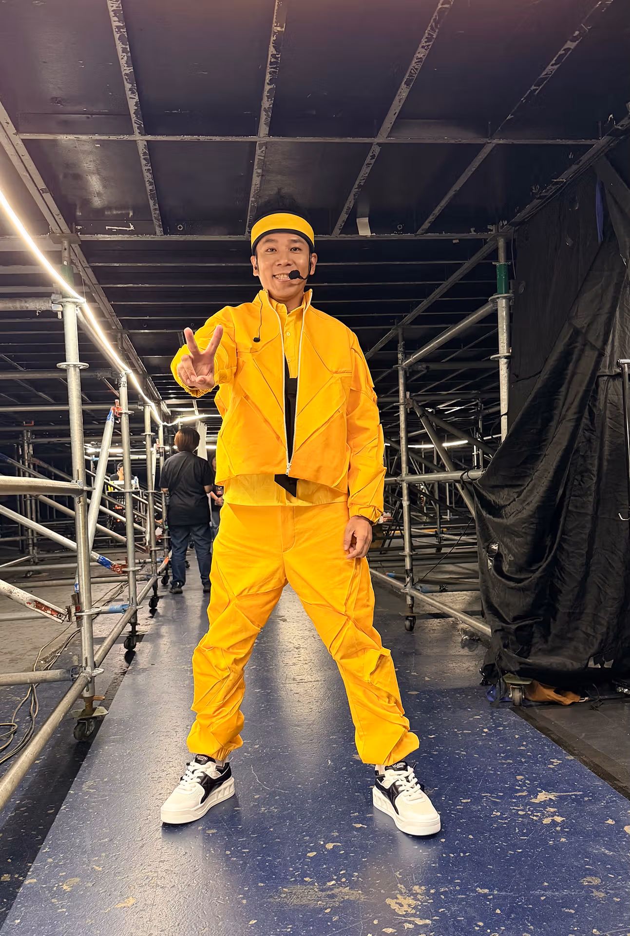 In an all-yellow outfit with headset microphone, Banana Brother makes a V sign among scaffolding and curtains