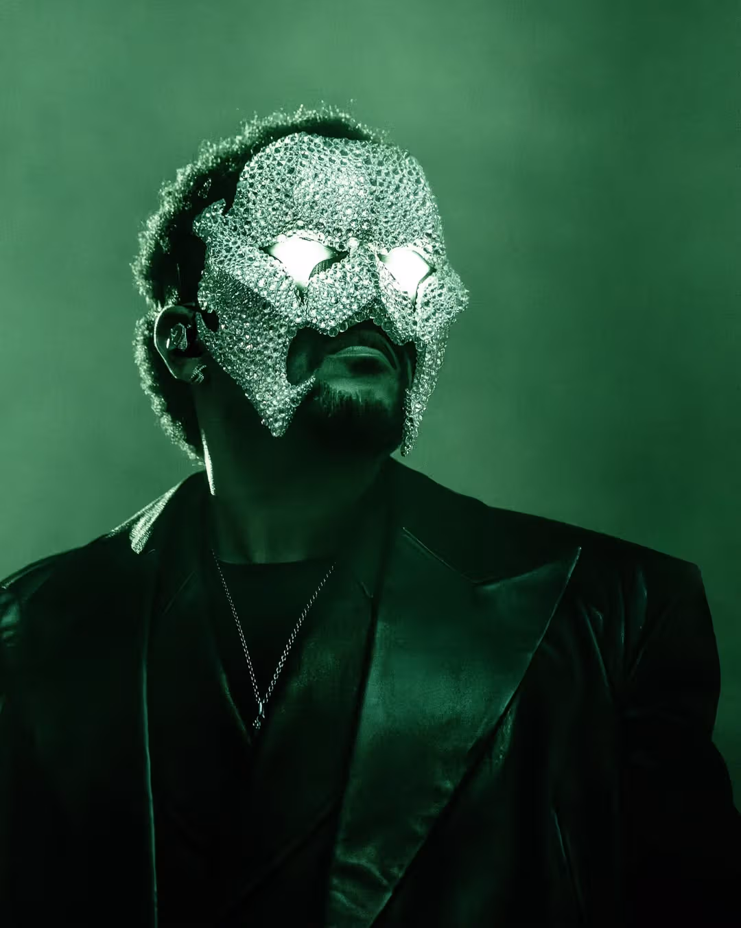 The Weeknd 戴银色质感面罩的近照，绿光落在脸侧与黑色衣领上。