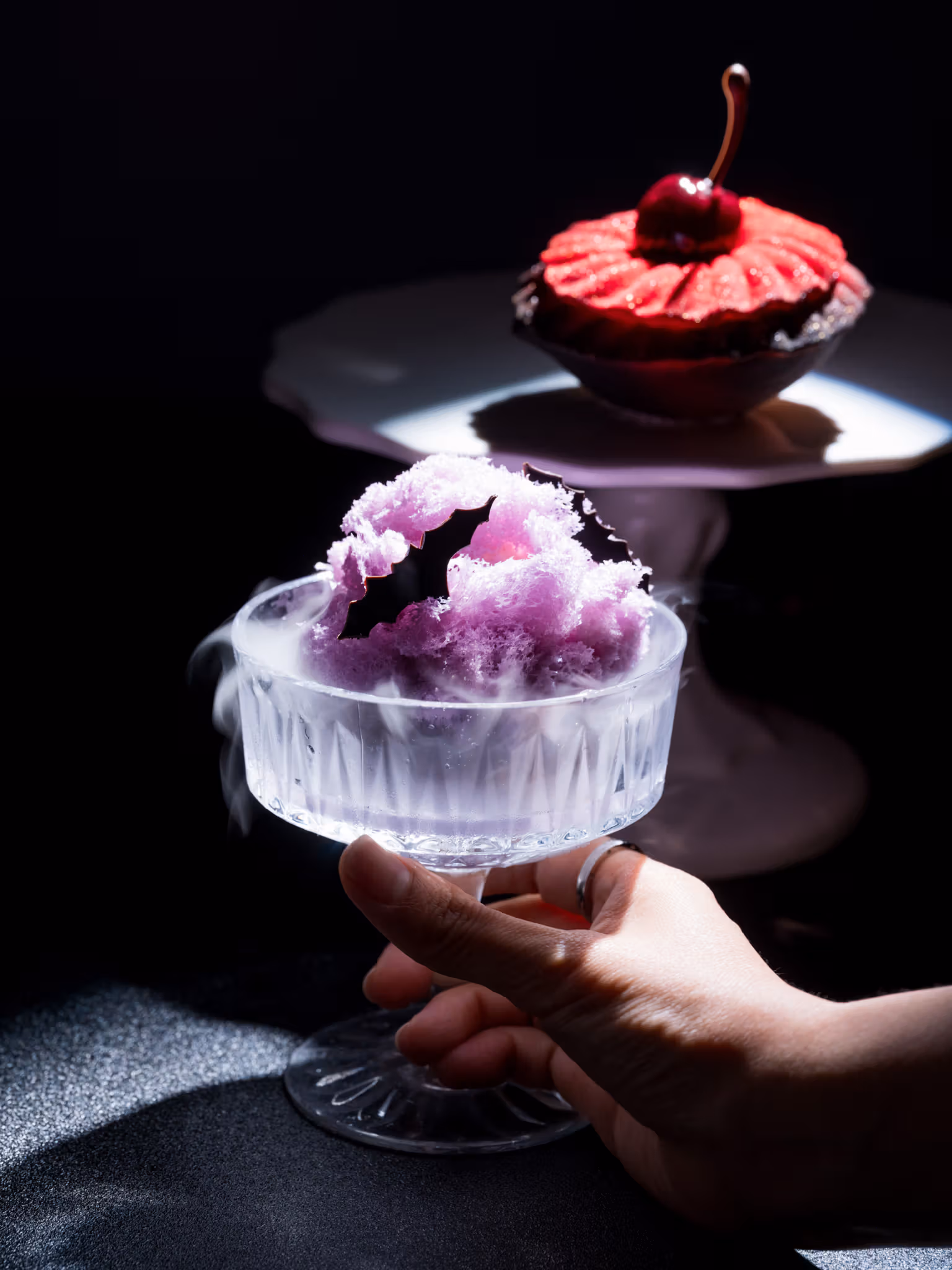 A hand holds a transparent stemmed vessel with a purple dessert, with a red cherry dessert behind