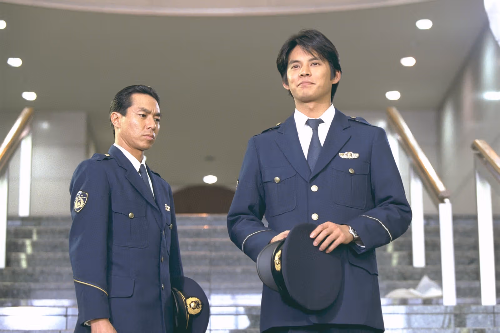 Two men in dark-blue uniforms stand indoors, one holding a cap
