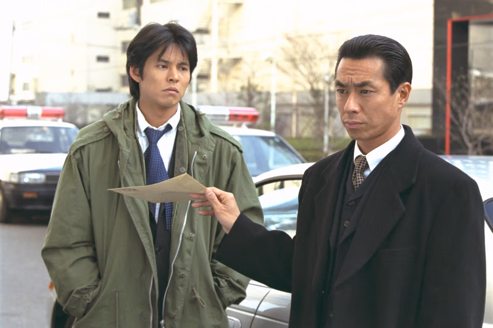 A man in a green coat stands beside a dark-suited man holding a document near a car