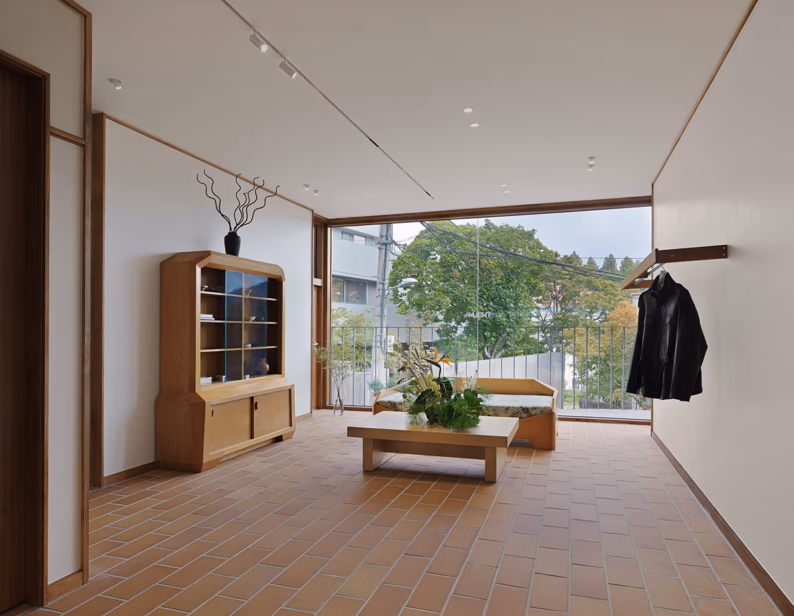A wooden display cabinet and low table beside a broad window overlooking trees and buildings