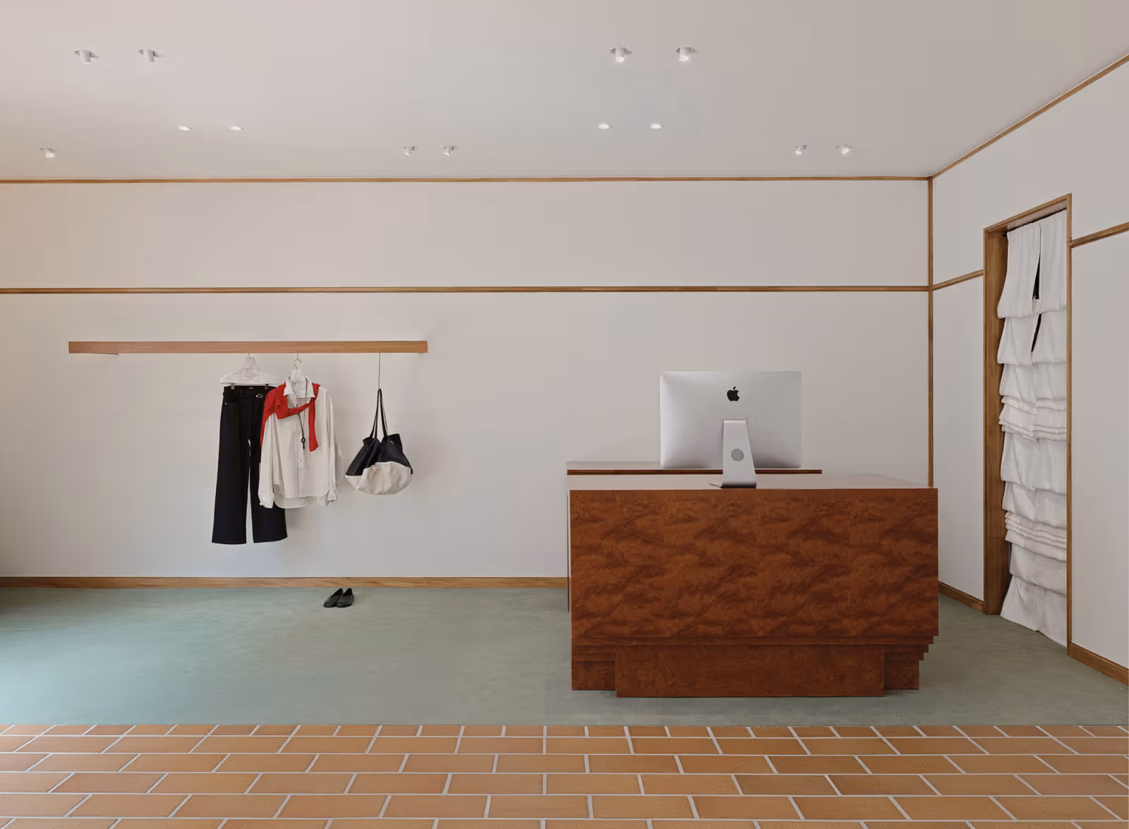 A computer on a wooden counter, clothes hanging to the left, and green and brick-coloured flooring