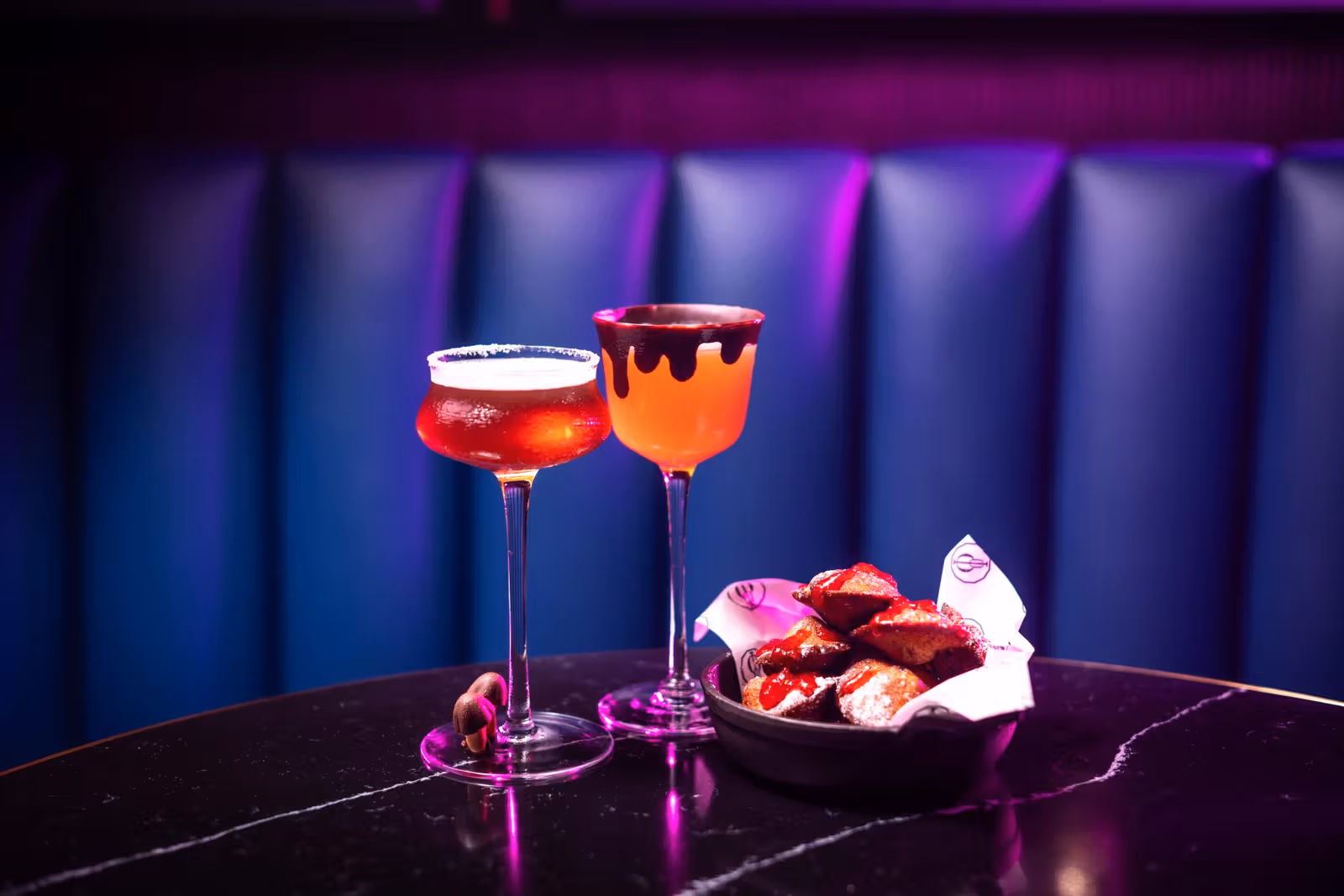 Two red and orange stemmed cocktails with a plate of snacks on a dark table