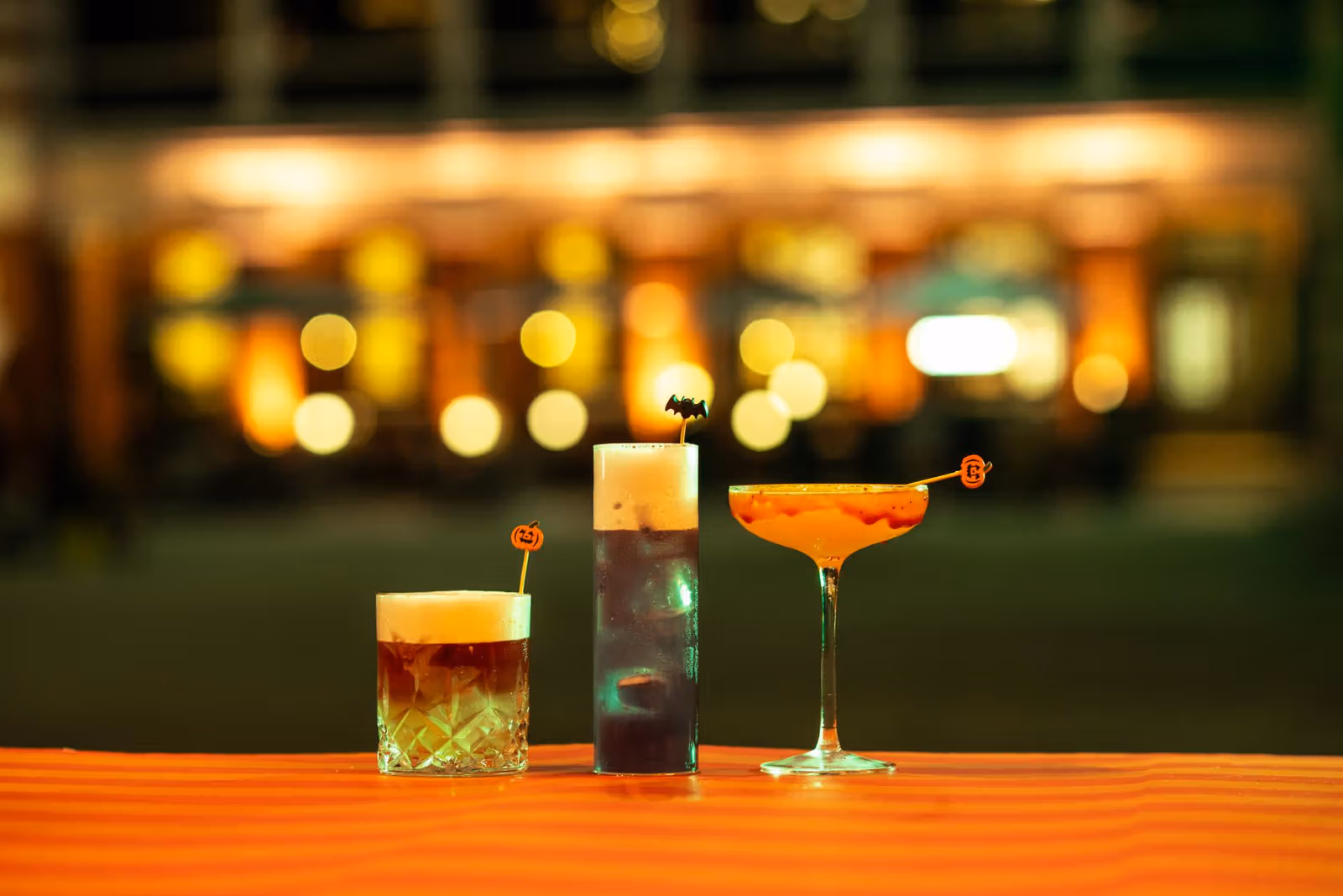 Three drinks of varying heights with bat and pumpkin decorations on a wooden bar