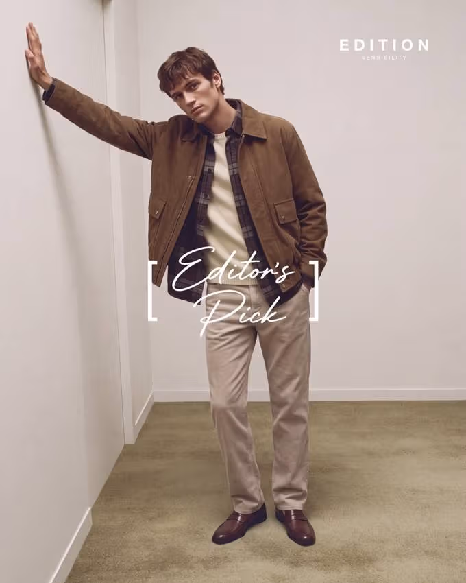 A standing model rests one hand on a wall, wearing a brown jacket, checked shirt and beige trousers.
