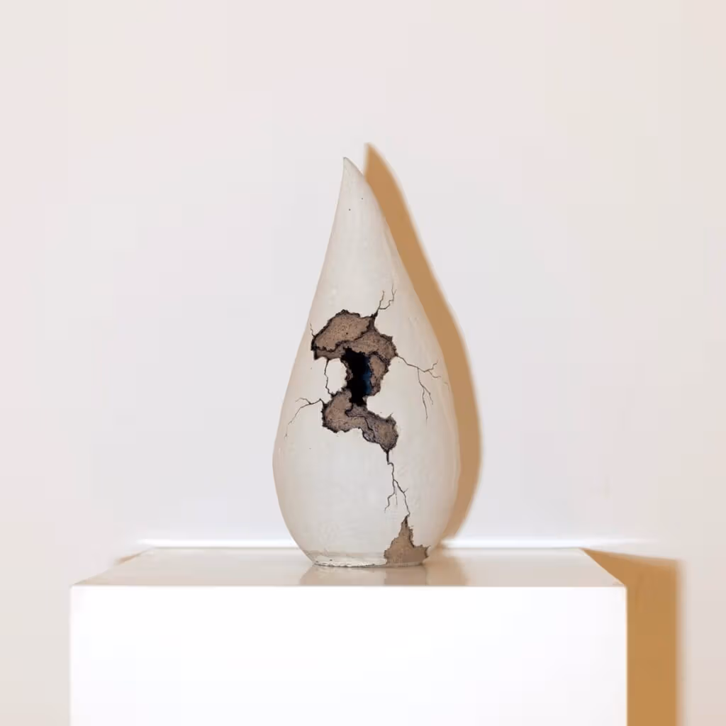 A pale, pointed, teardrop-shaped ceramic stands on a white plinth, with a dark opening and branching cracks across its middle.