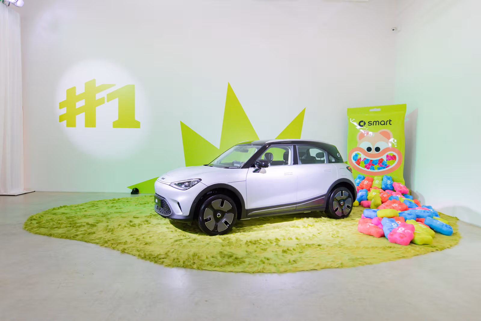 smart #1 สีขาวจอดบนพรมขนยาวสีเขียว ข้างรถมีวัตถุรูปหมีหลากสีกับฉากทรงบรรจุภัณฑ์พิมพ์คำว่า smart