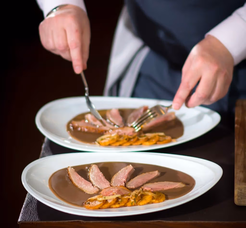 A server adjusts sliced meat on a white plate using a spoon and fork; both plates contain brown sauce.