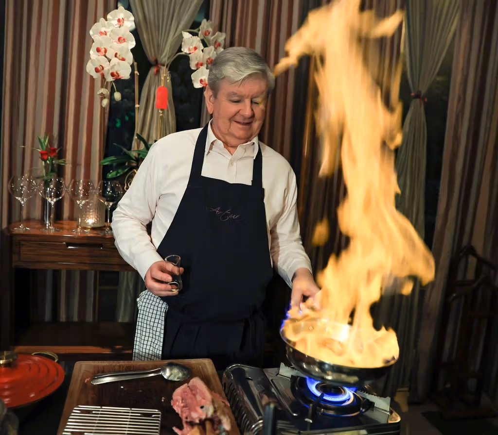 A server in a white shirt and dark apron stands beside a trolley as flames rise from a frying pan.