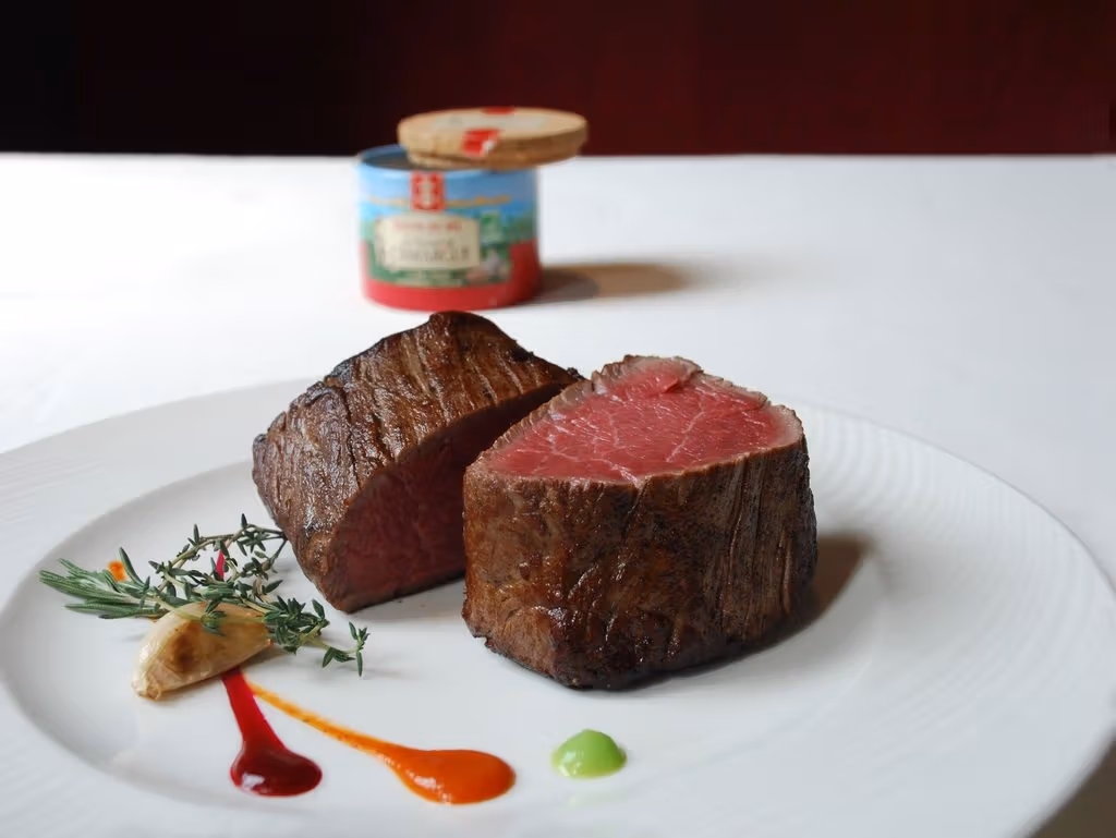 Two thick pieces of cut steak with red centres sit on a white plate with herbs, garlic and small amounts of coloured sauce.