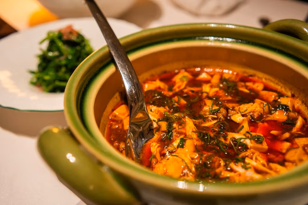 Tofu cubes, red sauce and spring-onion rings fill a green casserole with a metal serving spoon.