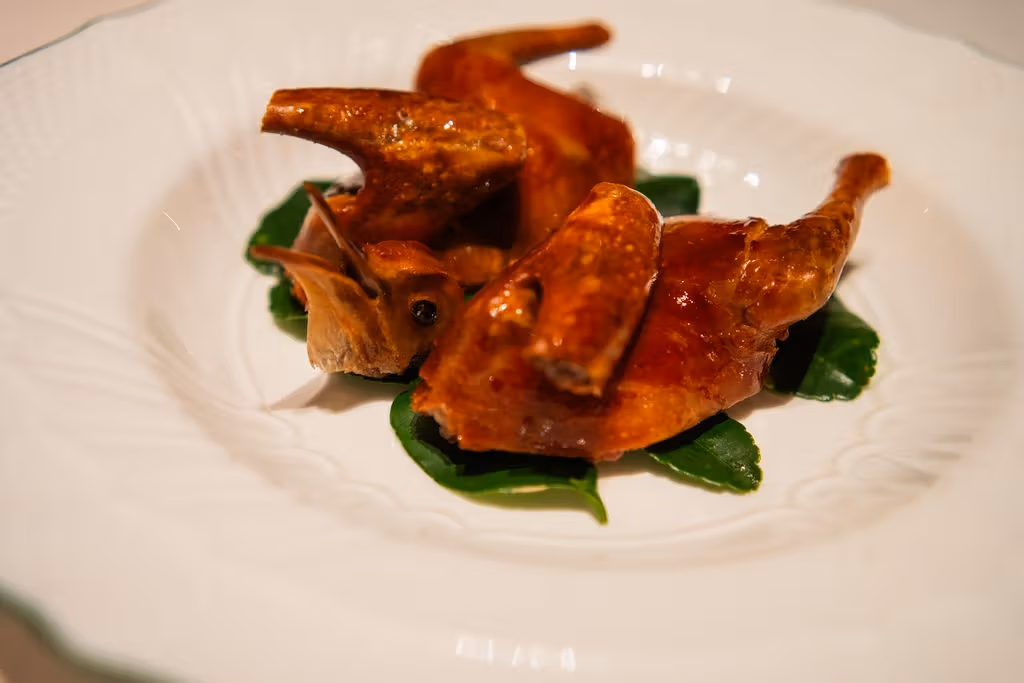 Pieces of brown-skinned pigeon sit on several dark-green leaves on a white plate.