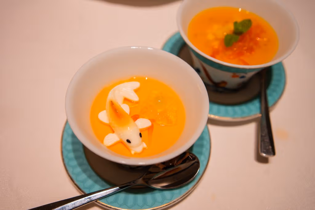 The front bowl contains a white fish-shaped pudding on an orange-yellow base; the dessert behind has diced fruit and mint leaves.