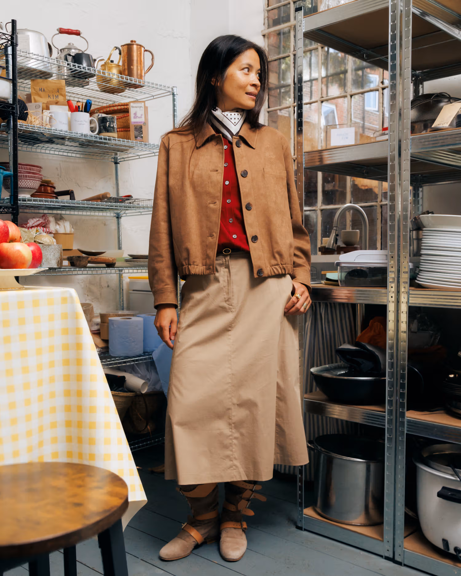 Kim in a brown cropped jacket, red layer and long skirt among metal shelves