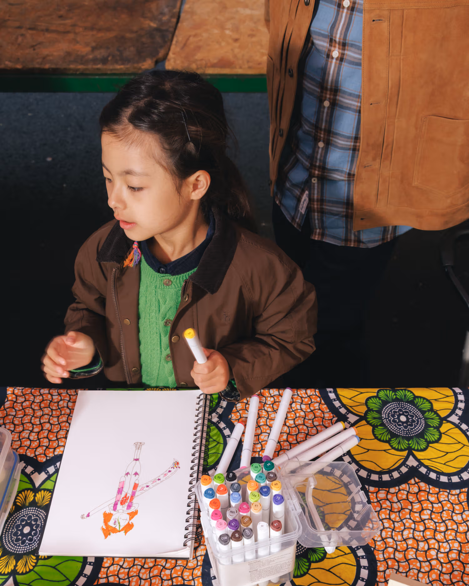 Child in a green knit and brown jacket drawing at a table