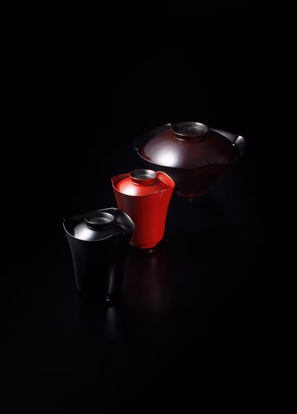 黒い背景に、起伏する縁と蓋を持つ黒、赤、暗褐色の漆器が三点並ぶ