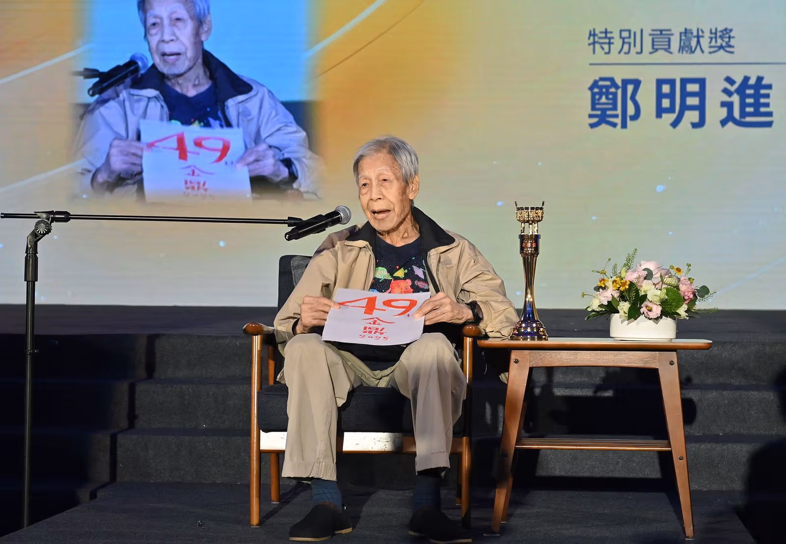 เจิ้งหมิงจิ้นนั่งหน้าไมโครโฟน ถือกระดาษ Golden Tripod ครั้งที่49 ปี2025 มีถ้วยข้างตัวและภาพฉายชื่อกับรางวัลคุณูปการพิเศษ